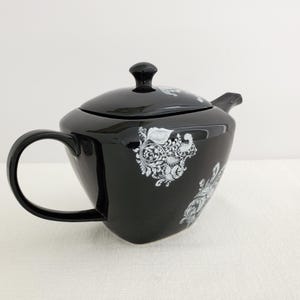 May include: A black ceramic teapot with a matching lid and handle. The teapot features a white floral design on the body and lid. The teapot is designed for brewing tea.