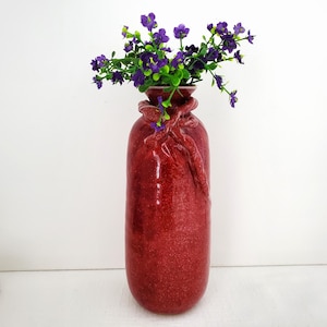 May include: A tall, red vase with a textured finish and a decorative knot design near the top. The vase contains a bouquet of purple flowers with green foliage. A decorative item for the home.