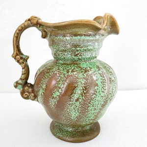 May include: A green and brown glazed ceramic pitcher with a ruffled rim and a decorative handle. The pitcher has a distressed look with a mottled glaze.