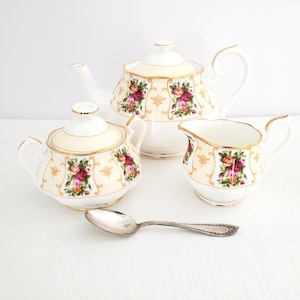 Puede incluir: Un juego de té vintage con tetera, azucarero y jarrita para la leche, todos con base crema y estampado floral. Los bordes están adornados con detalles dorados. Una cuchara plateada descansa frente al conjunto.