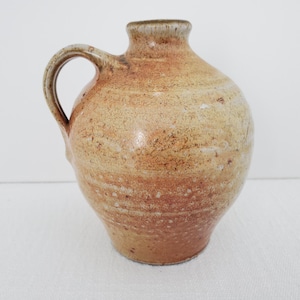 May include: A small, round, brown ceramic vase with a handle. The vase has a light brown glaze with a speckled finish.