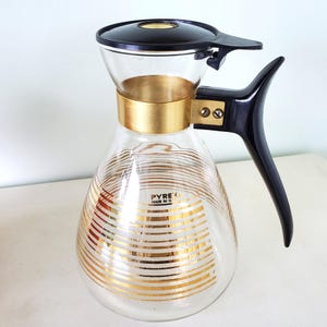 May include: A clear glass coffee pot with a gold and white striped design. The pot has a black plastic handle and lid. The label on the pot reads "Pyrex Made in U.S.A."
