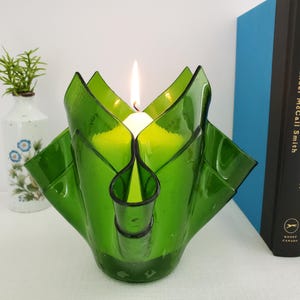 May include: A green glass candle holder with a lit white candle inside. The candle holder has a unique, petal-like design. A small vase with greenery and a book are in the background.