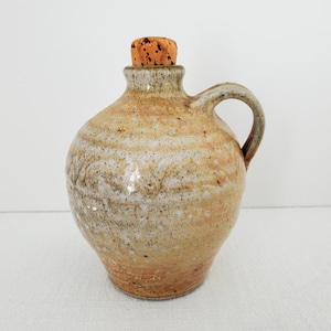 Op de afbeelding: Een handgemaakte keramische kan met een kurken stop. De kan is geglazuurd in een lichtbruine en witte kleur met een gespikkelde afwerking.