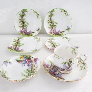 以下が含まれることがあります： 花柄のヴィンテージティーカップとソーサーのセット。カップとソーサーは白地に緑色の縁取りが施され、木と花の絵が描かれています。