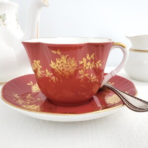 May include: A red teacup and saucer with a gold floral pattern. The cup has a scalloped rim and a delicate handle. The saucer has a matching pattern and a gold rim.