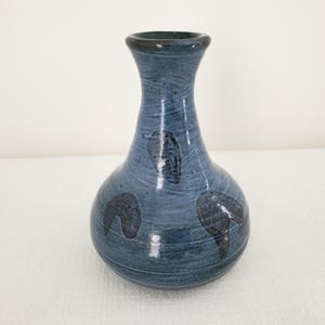Peut inclure: Un vase en céramique avec un vernis bleu brillant et une finition texturée sombre et tourbillonnante. Le vase présente une base large et arrondie qui se rétrécit vers un col étroit et un bord évasé. Trois motifs sombres en forme de feuille.