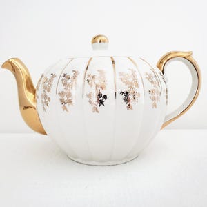 Puede incluir: Tetera de porcelana blanca con detalles dorados. La tetera tiene una forma redondeada con estrías verticales y motivos florales dorados. El pico, el asa y la tapa son dorados. Una pieza clásica para tomar el té o para exhibir.