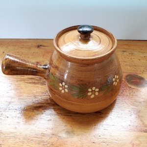May include: A brown ceramic pot with a lid and a handle. The pot is decorated with a green and white floral design.