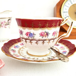 Può includere: Un set da tè composto da una tazza e un piattino, di colore rosso e bianco, con un bordo dorato e un motivo floreale. La tazza ha un manico delicato e il piattino ha un bordo ondulato.