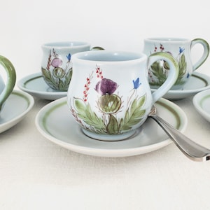 Könnte beinhalten: Ein Set hellblauer Keramik-Tassen und Untertassen. Jede Tasse zeigt ein handgemaltes Blumendesign mit lila Disteln, grünen Blättern und roten Akzenten. Die Henkel und Ränder sind grün. Ein silberner Löffel liegt auf einer Untertasse.