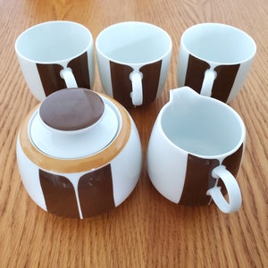 Op de afbeelding: Een set van bruine en witte keramische theekopjes met een bijpassende roomkan en suikerpot. De kopjes hebben een bruin streepontwerp.