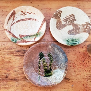 May include: Three round ceramic plates with textured surfaces. The top left plate is cream with brown and green brushstroke patterns. The top right plate is off-white with brown landscape-style designs. The bottom plate is dark brown with green accents.