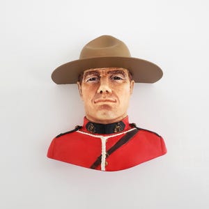Könnte beinhalten: Eine Wandbüste aus Keramik, die einen Beamten der Royal Canadian Mounted Police darstellt. Die Figur trägt einen beigefarbenen Hut mit breiter Krempe und eine leuchtend rote Uniformjacke mit schwarzem Kragen, goldenen Knöpfen und weißer Kordel.