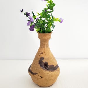 Puede incluir: Un jarrón de cerámica marrón con un diseño de espiral y un cuello estrecho. El jarrón está lleno de flores moradas y hojas verdes.