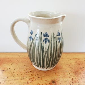 May include: A ceramic pitcher with a cream-coloured base and a speckled finish. The exterior features hand-painted green grass blades and small blue floral accents. It has a curved handle on the left side and a wide, slightly flared pouring spout.