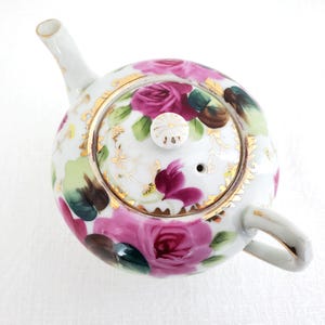 Può includere: Una teiera in porcellana bianca con coperchio, manico e beccuccio. La teiera è decorata con rose rosa, foglie verdi e dettagli dorati. Il coperchio ha un piccolo pomello bianco. La teiera è in stile vintage.