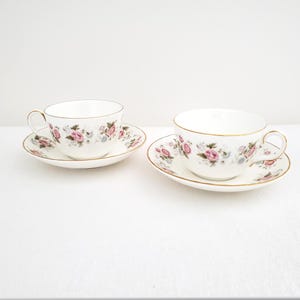 May include: Two white porcelain teacups and matching saucers featuring a delicate floral pattern of pink roses and small blue flowers. Each cup and saucer is finished with a thin gold trim along the rims and handles, creating a cohesive set.