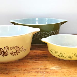 May include: Three vintage Pyrex mixing bowls in different sizes and colours. The largest bowl is green with a white floral pattern, the medium bowl is yellow with a brown floral pattern, and the smallest bowl is yellow with a green floral pattern.