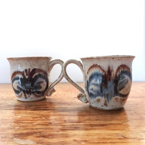 Peut inclure: Deux tasses en céramique avec un émail crème moucheté et un motif abstrait peint à la main dans des tons de brun et de bleu. Chaque tasse présente une anse incurvée et un bord évasé, révélant une finition rustique avec une texture d'argile.