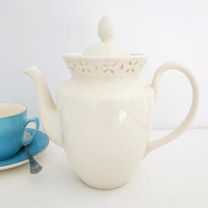 May include: A cream-coloured ceramic teapot with a decorative floral pattern around the rim and a matching lid. A blue teacup and saucer with a silver spoon are in the foreground. The teapot has a curved handle and spout.