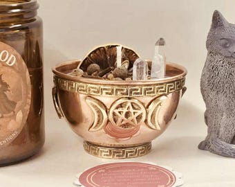 May include: An amber glass jar candle with a black lid, a copper-colored bowl with a pentagram design, and a gray cat-shaped candle. The jar has a label that reads "Sisterhood Candles with Intention."