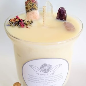 May include: A large, cream-colored candle with the text "Love Spell" on the label. The candle contains crystals, dried flowers, and a small jar with a cork stopper. The candle is surrounded by crystals and dried herbs.