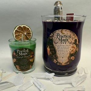 May include: A green candle in a glass jar topped with a dried lime slice and a larger purple candle in a glass jar featuring a label that reads Practical Magic, decorated with a small glass vial of herbs and a miniature book on top.