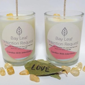 May include: Two glass jar candles with the text "Bay Leaf Intention Request" on a white label. The candles are surrounded by yellow crystals and bay leaves, one of which has the word "LOVE" written on it. Candles With Intentions.