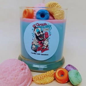 May include: A layered candle with blue and pink wax, decorated with a blue donut, orange donut, and yellow pastry. The candle has a label with the text "Captain Spaulding's Ice Cream" and a cartoon image. Additional decorative items include a pink scoop and other colorful treats.