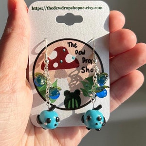 May include: A pair of silver dangle earrings with blue and green glass beads shaped like blueberries. The earrings are hanging from a card with the text "The Dew Drop Shop" and a mushroom illustration.