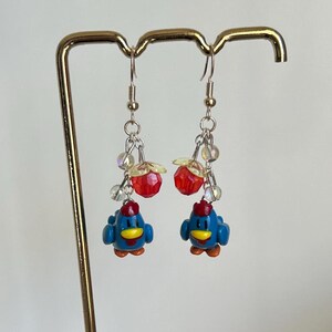 May include: A pair of dangle earrings featuring blue chicken charms with yellow beaks and red accents. The charms are attached to silver chains with clear and red beads.