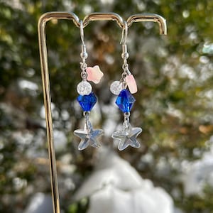 May include: Dangling earrings with a celestial theme. Each earring features a silver-tone hook, a cluster of beads, a blue diamond-shaped bead, and a clear star charm. Pink star and rectangle accents add color to the design. The earrings are displayed on a gold-colored stand.