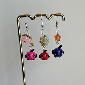 May include: Three pairs of dangle earrings with a variety of charms. The earrings feature a pink flower, a green leaf, and a gold star. The charms are attached to a small, round, colorful bead.