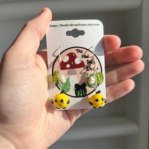 May include: A pair of dangle earrings featuring yellow and black bee charms with green and white flower accents. The earrings are on a white card with the text "The Dew Drop Shop" and a website address.
