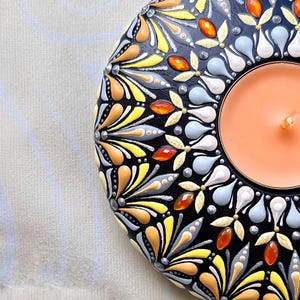 May include: A round, black candle holder with a detailed, colorful floral design. The design includes yellow, orange, and gray accents. A lit orange candle sits in the center of the holder, creating a warm glow.
