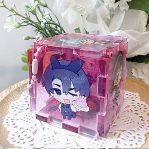 May include: A pink, transparent, cube-shaped display case with a cartoon character inside. The case has a hinged lid and a small pink chain. The character is holding a pink plate with a small pink cake. The text "Heikupon" is visible.