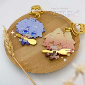 May include: Two anime-style character keychains. One has blue hair and holds a golden spoon, the other has pink hair and holds a golden fork. Both are on a wooden tray with a wheat stalk. The keychains have gold-colored attachments.
