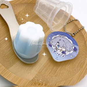 May include: A light blue and white dessert in a spoon, with a clear plastic container and a keychain with a cartoon character on it. The dessert is on a wooden tray.