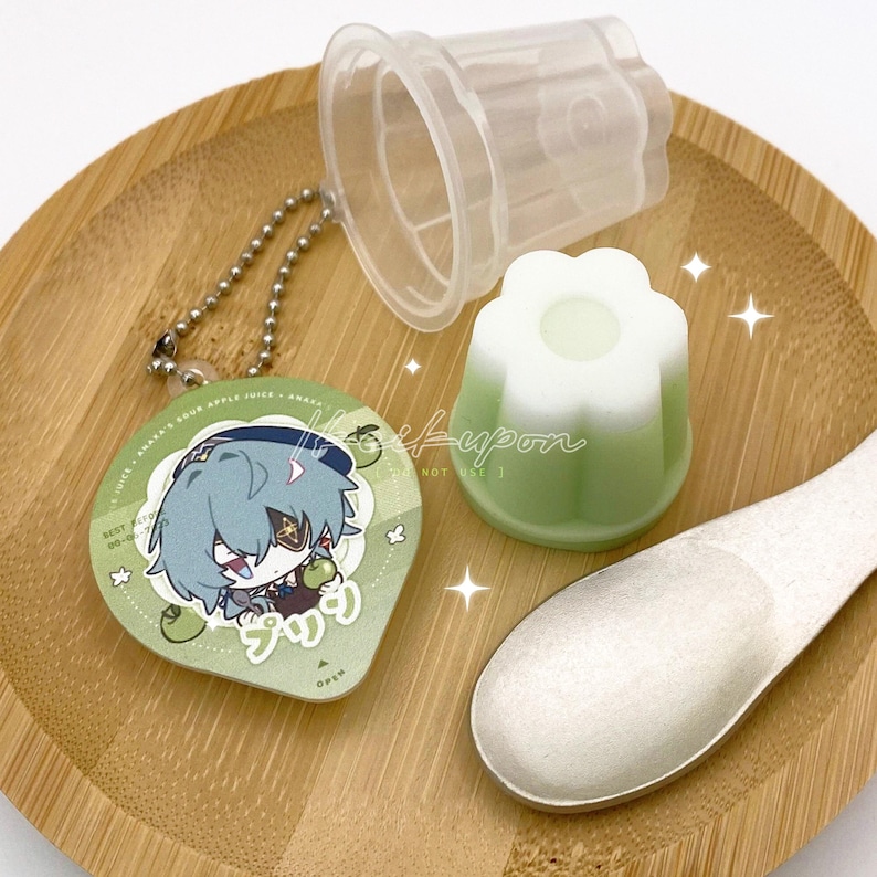 May include: A collection of items including a green and white flower-shaped dessert, a clear plastic container, a spoon, and a keychain with a cartoon character illustration. The items are arranged on a wooden tray.