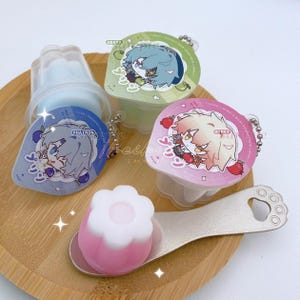May include: A collection of three colorful, round, dessert containers with character illustrations and a pink and white flower-shaped dessert on a spoon. The containers are blue, green, and pink, with silver keychains attached. The spoon has a paw print design.