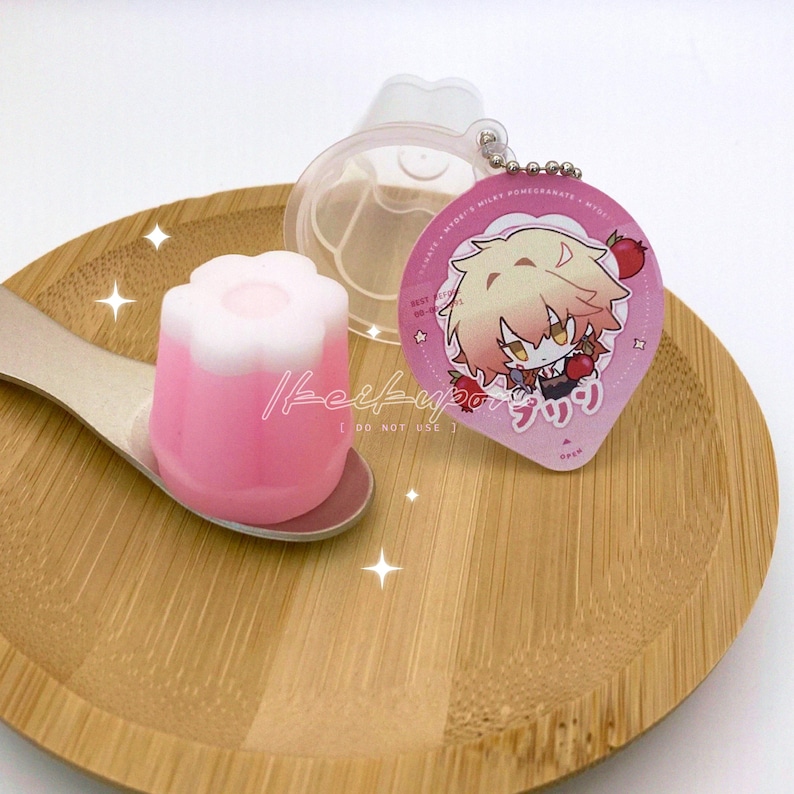 May include: A pink and white dessert in a flower shape sits on a wooden spoon. A clear plastic container and a pink keychain with a cartoon character and Japanese text are also present. The items are on a wooden plate.