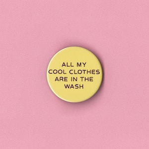 May include: A yellow button with the text "ALL MY COOL CLOTHES ARE IN THE WASH" in black, centered on a pink background. The button is circular and has a glossy finish, with the text in a simple, sans-serif font.