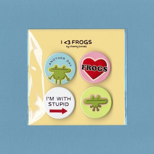 May include: A pack of four circular pin badges with frog illustrations and text. The badges are packaged on a yellow card. Text includes "I <3 FROGS", "Another Day", "FROGS", and "I'M WITH STUPID".