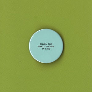 May include: A circular light blue pinback button featuring the black text "ENJOY THE SMALL THINGS IN LIFE" centered on its surface. The object has a smooth, matte finish and a clean, simple design with no additional decorative elements or patterns.