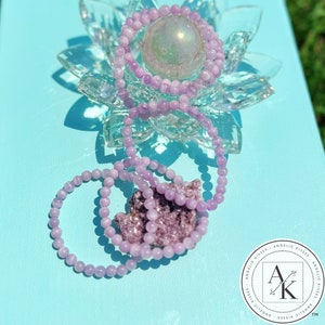 May include: A collection of light purple beaded bracelets and a cluster of amethyst crystals are displayed on a light blue surface. The bracelets are draped over a clear crystal lotus flower and a spherical iridescent object.