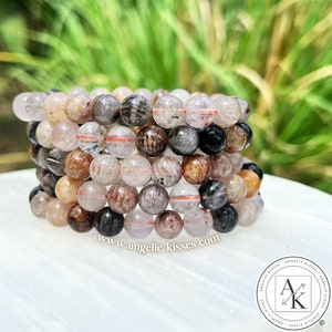 May include: Stack of gemstone bracelets featuring a variety of colors including clear, brown, black, and peach. The beads are round and strung together. The website address www.angelic-kisses.com is visible at the bottom.
