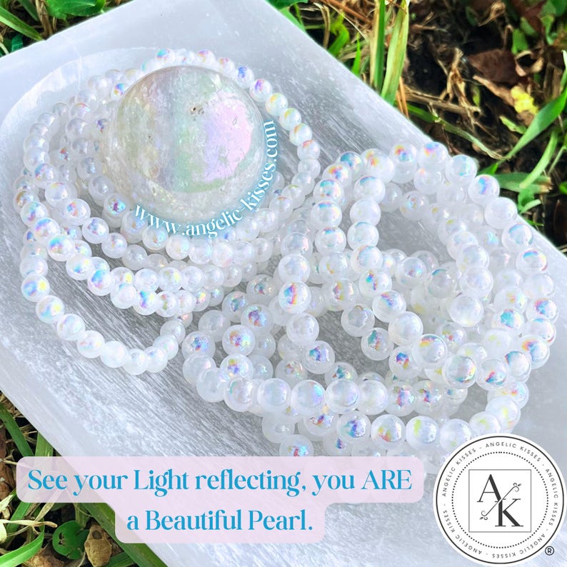 Op de afbeelding: Meerdere witte kralenarmbanden en een grote iriserende bol liggen op een wit oppervlak. De armbanden zijn in een cirkelvorm gerangschikt. De afbeelding bevat de tekst "See your Light reflecting, you ARE a Beautiful Pearl."