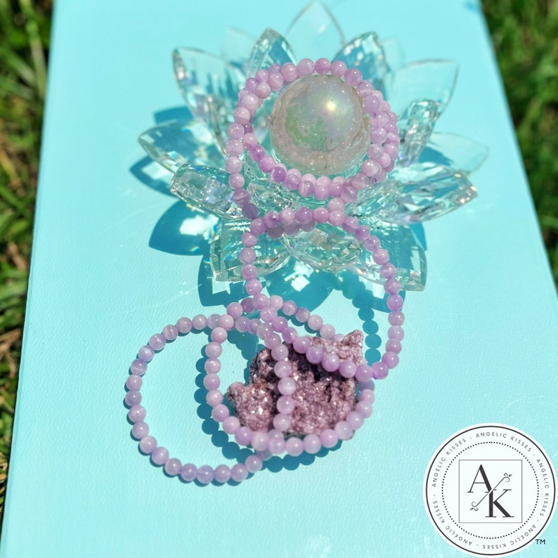 May include: A collection of light purple beaded bracelets and a cluster of amethyst crystals are displayed on a light blue surface. The bracelets are draped over a clear crystal lotus flower and a spherical iridescent object.