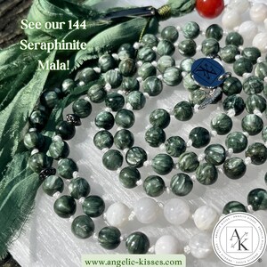 Op de afbeelding: Een mala-ketting met donkergroene seraphinietkralen met witte vezelige insluitingen, afgewisseld met gladde witte kralen en zilverkleurige metalen accenten. Een blauwe ronde bedel met logo en een rode kraal zijn bevestigd. Tekst: See our 144 Seraphinite Mala! en www.angelic-kisses.com.